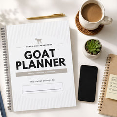 A spiral-bound 'GOAT PLANNER' for herd and kid management is displayed in a styled flat-lay on a white surface, surrounded by a cup of coffee, a pen, a small succulent, a smartphone, and another notebook.