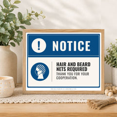 A framed printed notice sign on a tabletop in a styled interior setting. The sign has a blue border, a blue header reading “NOTICE,” and text stating “HAIR AND BEARD NETS REQUIRED THANK YOU FOR YOUR COOPERATION.