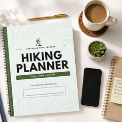 A spiral-bound hiking planner titled 'Exploring Trail Tracker Hiking Planner' shown in a styled flat-lay with a cup of coffee, a pen, a small succulent, a smartphone, and another notebook on a white surface.