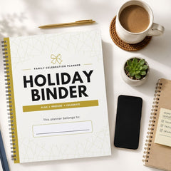A spiral-bound 'Holiday Binder' family celebration planner is displayed in a styled flat-lay arrangement on a white surface, surrounded by a cup of coffee, a smartphone, a small succulent, and other notebooks.