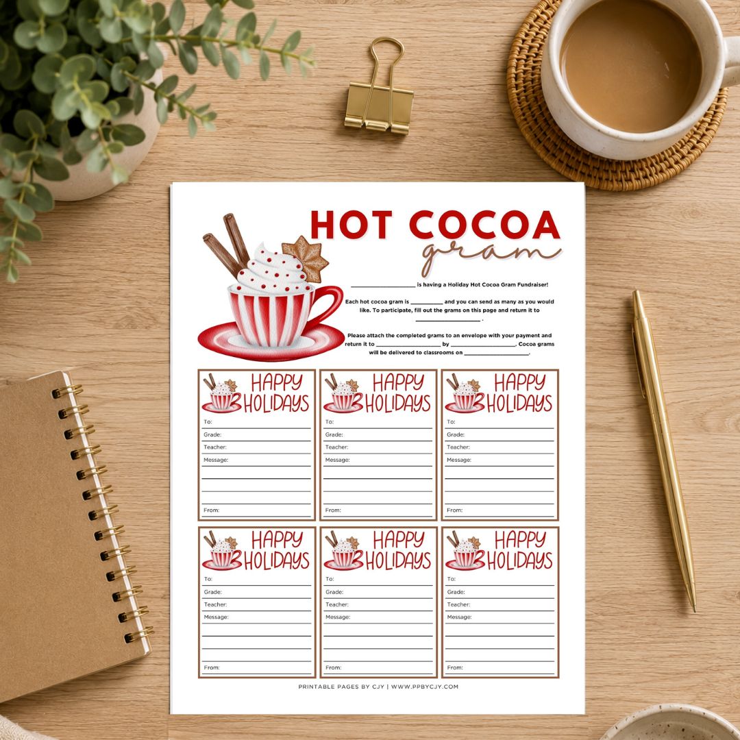 A printable hot cocoa gram worksheet sheet displayed on a wooden desk with a coffee cup, plant, notebook, and pen around it. The sheet features a festive red and white hot cocoa graphic and multiple 'Happy Holidays' writing sections.