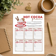 A printable hot cocoa gram worksheet sheet displayed on a wooden desk with a coffee cup, plant, notebook, and pen around it. The sheet features a festive red and white hot cocoa graphic and multiple 'Happy Holidays' writing sections.