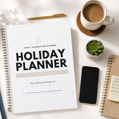 A spiral-bound holiday planner notebook lies open on a white desk, surrounded by a coffee cup on a coaster, a small succulent, a smartphone, and another notebook. The planner cover reads 'HOLIDAY PLANNER' with 'Family Celebration Planner' above it.