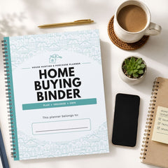 A spiral-bound 'Home Buying Binder' planner is displayed in a styled flat-lay arrangement on a white surface, surrounded by a coffee cup, a pen, a small succulent, a smartphone, and another notebook.