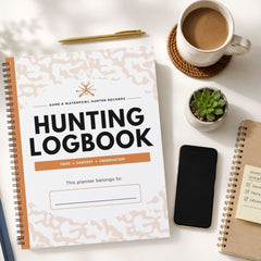 A spiral-bound hunting logbook with a white cover featuring a light beige camouflage pattern and bold black text, styled in a flat-lay arrangement on a white desk alongside a coffee cup, pen, phone, and other notebooks.