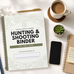 A spiral-bound 'Hunting & Shooting Binder' planner lies on a white desk surrounded by lifestyle props, including a cup of coffee, a pen, a smartphone, a small succulent, and another notebook.
