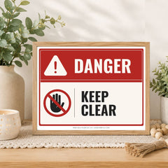 A framed warning sign with a red and white background, featuring the word 'DANGER' and a 'KEEP CLEAR' message with a crossed-out hand symbol. The sign is displayed on a wooden surface with decorative plants and candles in the background.