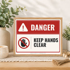 A framed warning sign on a tabletop in a styled home setting. The sign has a red and white background with the word 'DANGER' and a crossed-out hand symbol with the text 'KEEP HANDS CLEAR.