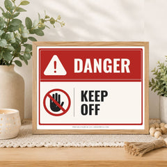 A framed warning sign with a red and white background, featuring the word 'DANGER' and a 'KEEP OFF' hand symbol, styled on a tabletop with plants and home decor.