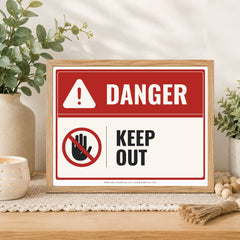 A framed warning sign with a red and white background, featuring the word 'DANGER' and a 'KEEP OUT' symbol, displayed on a tabletop with plants and home decor around it.