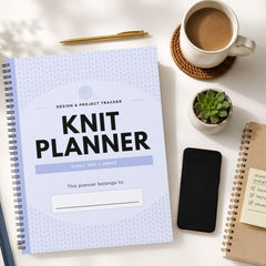 A spiral-bound 'Knit Planner' with a light blue and white chevron-patterned cover is displayed in a styled flat-lay arrangement on a white desk, surrounded by a coffee cup, a pen, a small succulent, a smartphone, and another notebook.