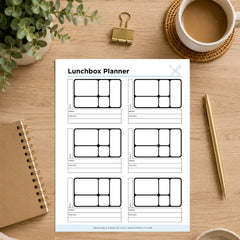 A printable lunchbox planner sheet laid on a wooden desk, surrounded by a coffee cup, binder clip, notebook, pen, and plant. The planner has six labeled slots for lunch, snack, drink, and notes, with a small butterfly logo in the top right.