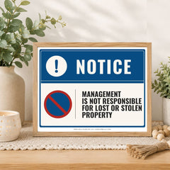 A framed warning sign with a blue and white background, featuring the word 'NOTICE' and a red-and-blue no symbol with the text 'Management is not responsible for lost or stolen property.' The sign is displayed on a tabletop in a styled interior setting with plants, a candle, and decorative beads.