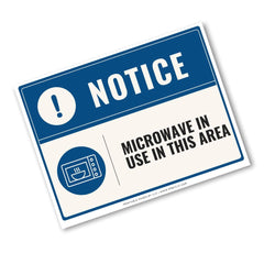 A rectangular warning sign with a blue header reading 'NOTICE' and a microwave icon, plus the text 'MICROWAVE IN USE IN THIS AREA' on an off-white background, shown at an angle on a white backdrop.