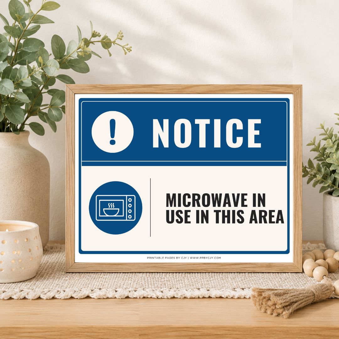 A framed rectangular warning sign on a tabletop in a styled interior setting. The sign reads “NOTICE” with a blue exclamation mark icon and “MICROWAVE IN USE IN THIS AREA” in black text.