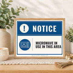 A framed rectangular warning sign on a tabletop in a styled interior setting. The sign reads “NOTICE” with a blue exclamation mark icon and “MICROWAVE IN USE IN THIS AREA” in black text.