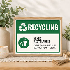 A framed recycling sign displayed on a wooden table in a styled home setting with plants and a candle. The sign features a green and white design with the text 'RECYCLING' and 'MIXED RECYCABLES THANK YOU FOR HELPING KEEP OUR PLANET CLEAN.