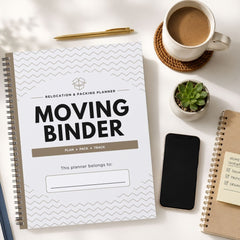 A spiral-bound moving binder planner lies on a white desk, surrounded by a coffee cup on a coaster, a small succulent, a smartphone, and another notebook. The planner cover is white with gray chevron lines and large black text reading 'MOVING BINDER'.