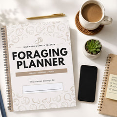 A spiral-bound Foraging Planner notebook is shown in a styled flat-lay arrangement on a white surface, surrounded by a coffee cup, pen, small succulent, smartphone, and another notebook.