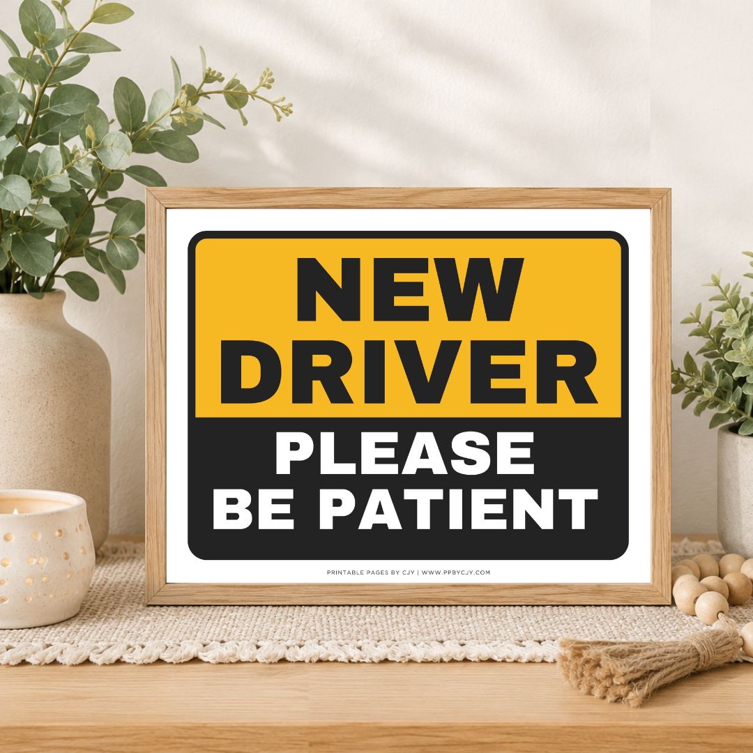 A framed novelty sign with the text 'NEW DRIVER PLEASE BE PATIENT' in bold black and white lettering on a yellow and black background, styled on a tabletop with plants and home decor.