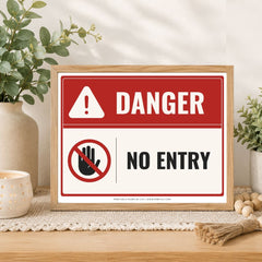 A framed warning sign with a red and white background, featuring the word 'DANGER' and a crossed-out hand symbol with the words 'NO ENTRY'. The sign is displayed on a wooden surface in a styled home setting with plants and decorative items.