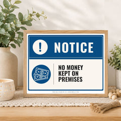 A framed novelty warning sign on a wooden table, styled with potted plants and a candle. The sign reads 'NOTICE' and 'NO MONEY KEPT ON PREMISES' with blue and white graphics.