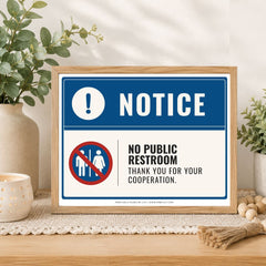 A framed novelty restroom sign displayed on a wooden table with decorative plants and candles. The sign has a blue header with a white exclamation mark and the word 'NOTICE,' and a white lower section with a red-and-blue no public restroom symbol and the text 'NO PUBLIC RESTROOM THANK YOU FOR YOUR COOPERATION.