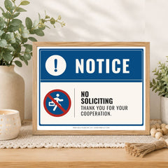 A framed novelty sign with a blue and white 'NOTICE' header and a red-and-white 'NO SOLICITING' symbol, displayed on a wooden surface with plants and home decor in the background.