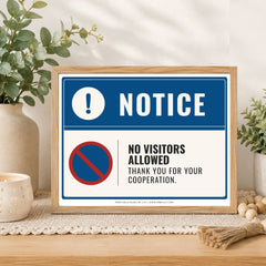 A framed novelty sign with a blue and white design that reads 'NOTICE' and 'NO VISITORS ALLOWED THANK YOU FOR YOUR COOPERATION.' The sign is displayed on a wooden table with decorative plants, a candle, and a burlap bundle in a styled indoor setting.