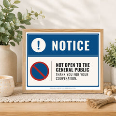 A framed printed notice sign on a tabletop, styled with plants and home decor. The sign has a blue header with a white exclamation mark and the word NOTICE, plus a red-and-blue no symbol and the text 'NOT OPEN TO THE GENERAL PUBLIC THANK YOU FOR YOUR COOPERATION.