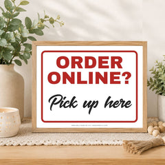 A framed sign on a wooden table with eucalyptus and decorative props in the background. The sign has a light wood frame, a white background, red border lines, and text reading 'ORDER ONLINE?' in red and 'Pick up here' in black.