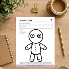A printable coloring page for a 'Voodoo Doll' shown in a styled flat-lay on a wooden desk, surrounded by a coffee cup, a notebook, a gold binder clip, and a plant.