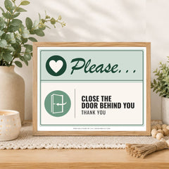 A framed printed sign on a tabletop in a styled interior setting. The sign has a light wood frame and reads 'Please.