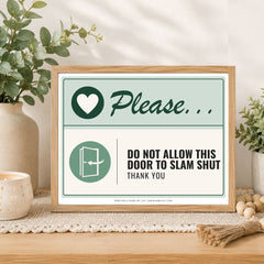 A framed decorative sign on a wooden table in a styled interior setting. The sign has a light wood frame and reads 'Please.
