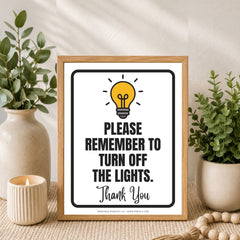 A framed typographic wall sign with a light bulb graphic and the text 'PLEASE REMEMBER TO TURN OFF THE LIGHTS. Thank You' sits on a tabletop styled with potted green plants, a candle, and a wooden bead runner.