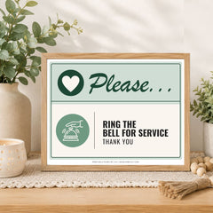 A framed printed sign on a tabletop in a styled interior setting. The sign has a light wood frame and reads 'Please.