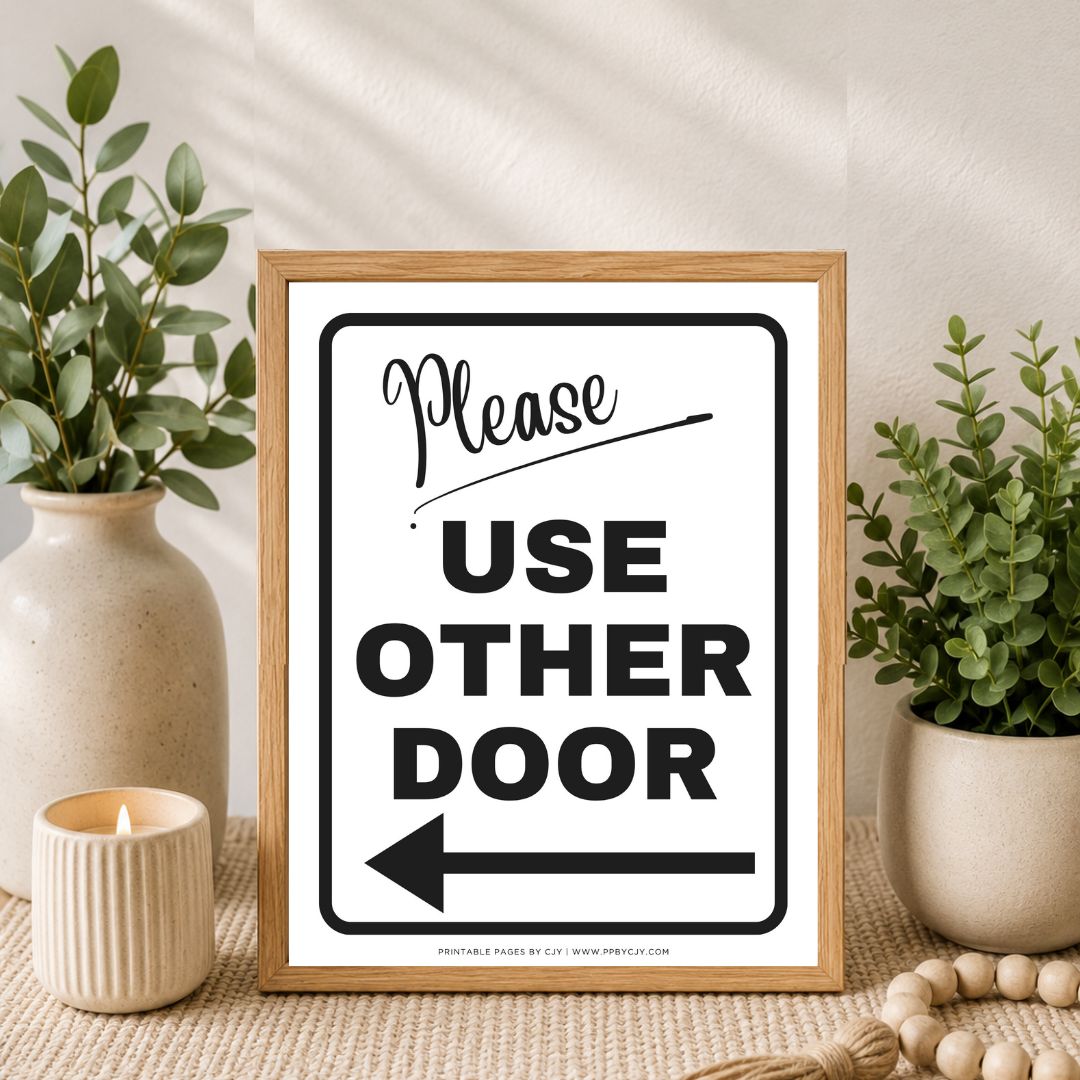 A framed 'Please USE OTHER DOOR' sign with a left-pointing arrow, styled on a tabletop with potted green plants, a candle, and a neutral wall background.