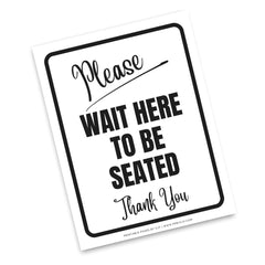 A white rectangular printed sign with a black border and black text reading 'Please WAIT HERE TO BE SEATED Thank You' on a white background.