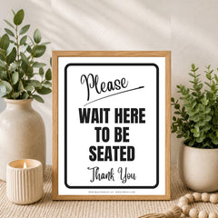 A framed printed sign reading 'Please WAIT HERE TO BE SEATED Thank You' in black text on a white background, styled in a home setting with a wooden frame, flanked by potted green plants and a lit candle.