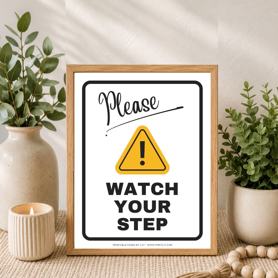 A framed 'Please Watch Your Step' warning sign with a yellow caution triangle, styled on a woven surface between two potted green plants and a small candle.
