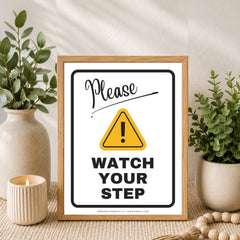 A framed 'Please Watch Your Step' warning sign with a yellow caution triangle, styled on a woven surface between two potted green plants and a small candle.