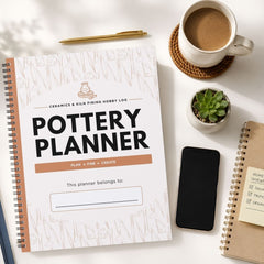 A spiral-bound Delivery Planner notebook is shown in a styled flat-lay arrangement on a white surface, alongside a cup of coffee, a pen, a small succulent, a smartphone, and another notebook.