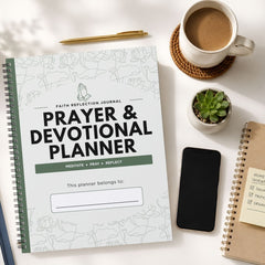 A spiral-bound Prayer & Devotional Planner journal is shown in a styled flat-lay arrangement on a white surface, alongside a cup of coffee, a pen, a small succulent, a smartphone, and another notebook.