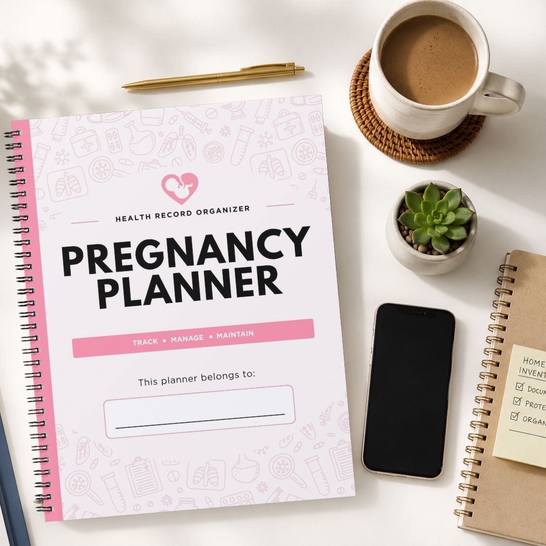 A spiral-bound pregnancy planner cover laid on a white surface, surrounded by a coffee cup on a coaster, a small succulent, a smartphone, and another notebook. The planner cover is white with pink line drawings and pink accents, and the title reads 'PREGNANCY PLANNER.