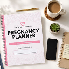 A spiral-bound pregnancy planner cover laid on a white surface, surrounded by a coffee cup on a coaster, a small succulent, a smartphone, and another notebook. The planner cover is white with pink line drawings and pink accents, and the title reads 'PREGNANCY PLANNER.