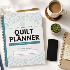 A spiral-bound quilt planner with a white cover and light blue geometric pattern, styled on a white desk with a coffee cup, pen, phone, and other notebooks.