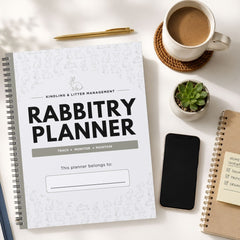 A spiral-bound Rabbitry Planner notebook is shown in a styled flat-lay arrangement on a white surface, surrounded by a cup of coffee, a small succulent, a smartphone, and another notebook.