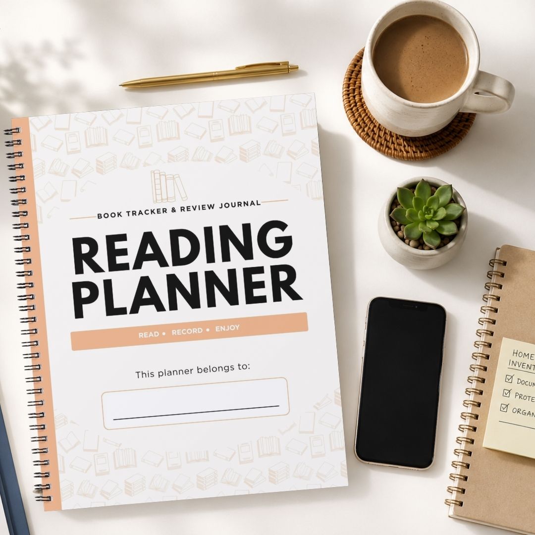 A spiral-bound 'Reading Planner' book tracker journal is displayed in a styled flat-lay arrangement on a white surface, surrounded by a cup of coffee, a smartphone, a small succulent, and other notebooks.