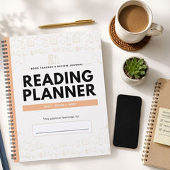 A spiral-bound 'Reading Planner' book tracker journal is displayed in a styled flat-lay arrangement on a white surface, surrounded by a cup of coffee, a smartphone, a small succulent, and other notebooks.