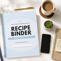 A spiral-bound recipe binder planner with a light blue and white gingham cover is displayed in a styled flat-lay on a white desk alongside a cup of coffee, a pen, a small succulent, a smartphone, and another notebook.